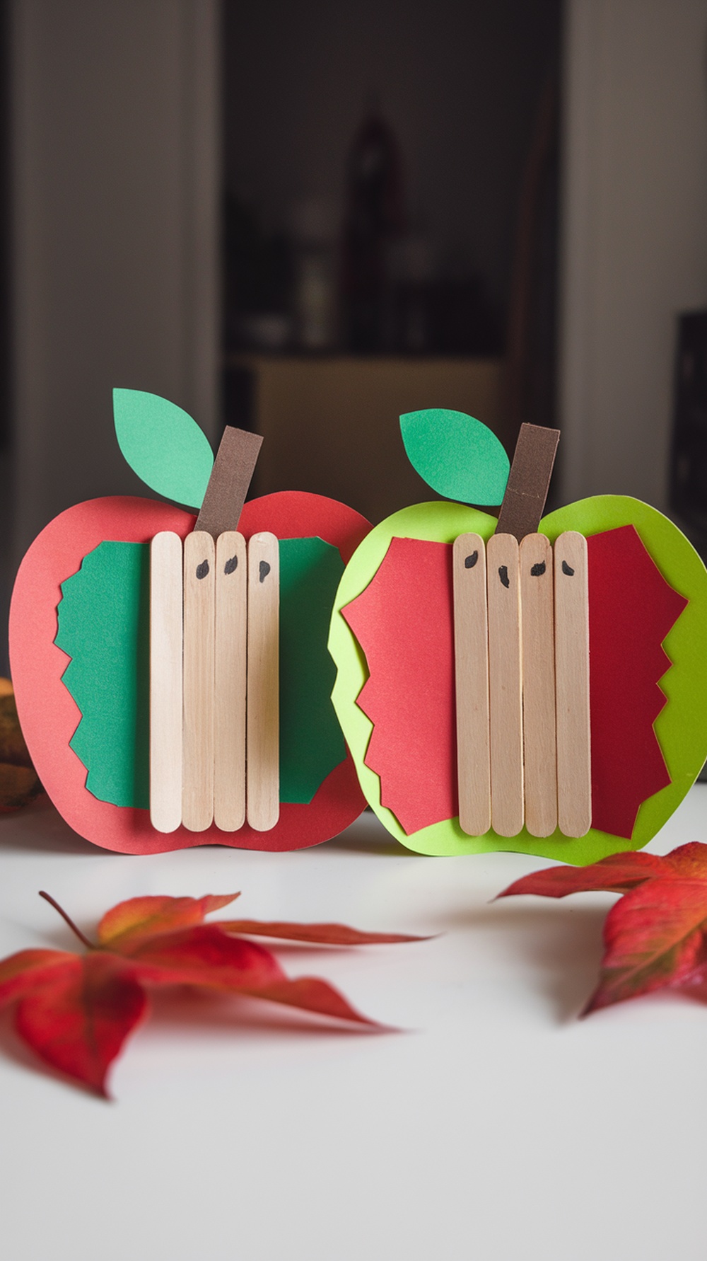 Colorful popsicle stick apple crafts in red and green on a flat surface with fall leaves