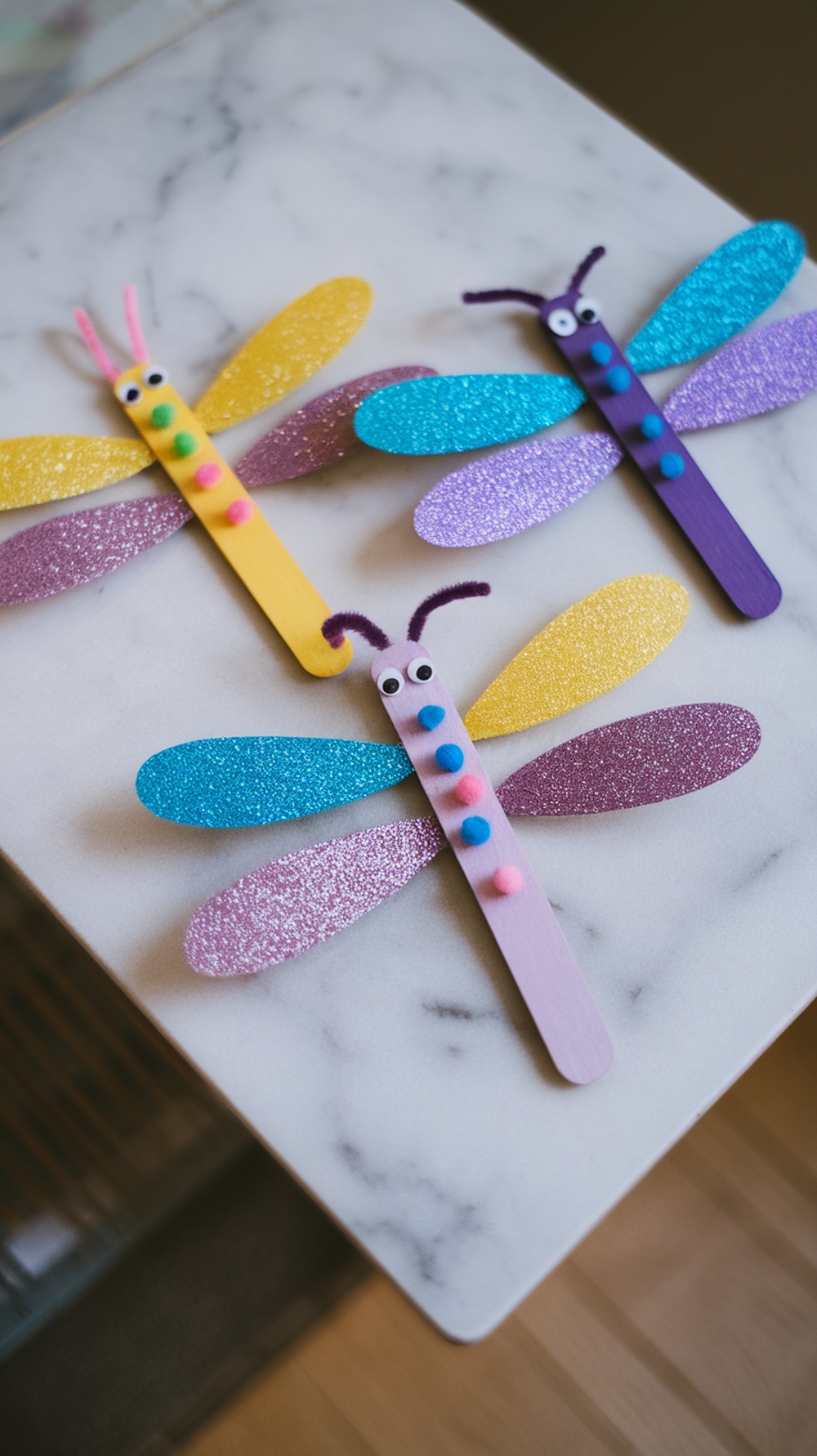 Colorful popsicle stick dragonflies with glittery wings on a marble surface.