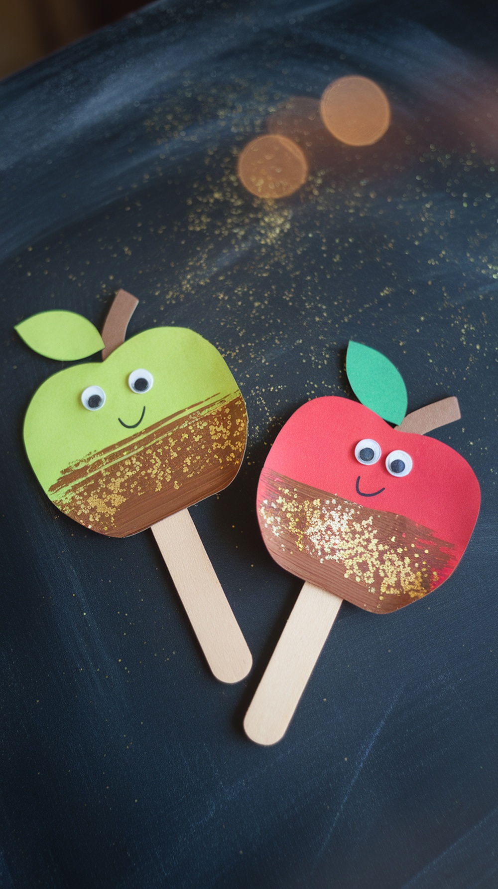 Two popsicle stick caramel apple puppets, one red and one green, with googly eyes and glitter.