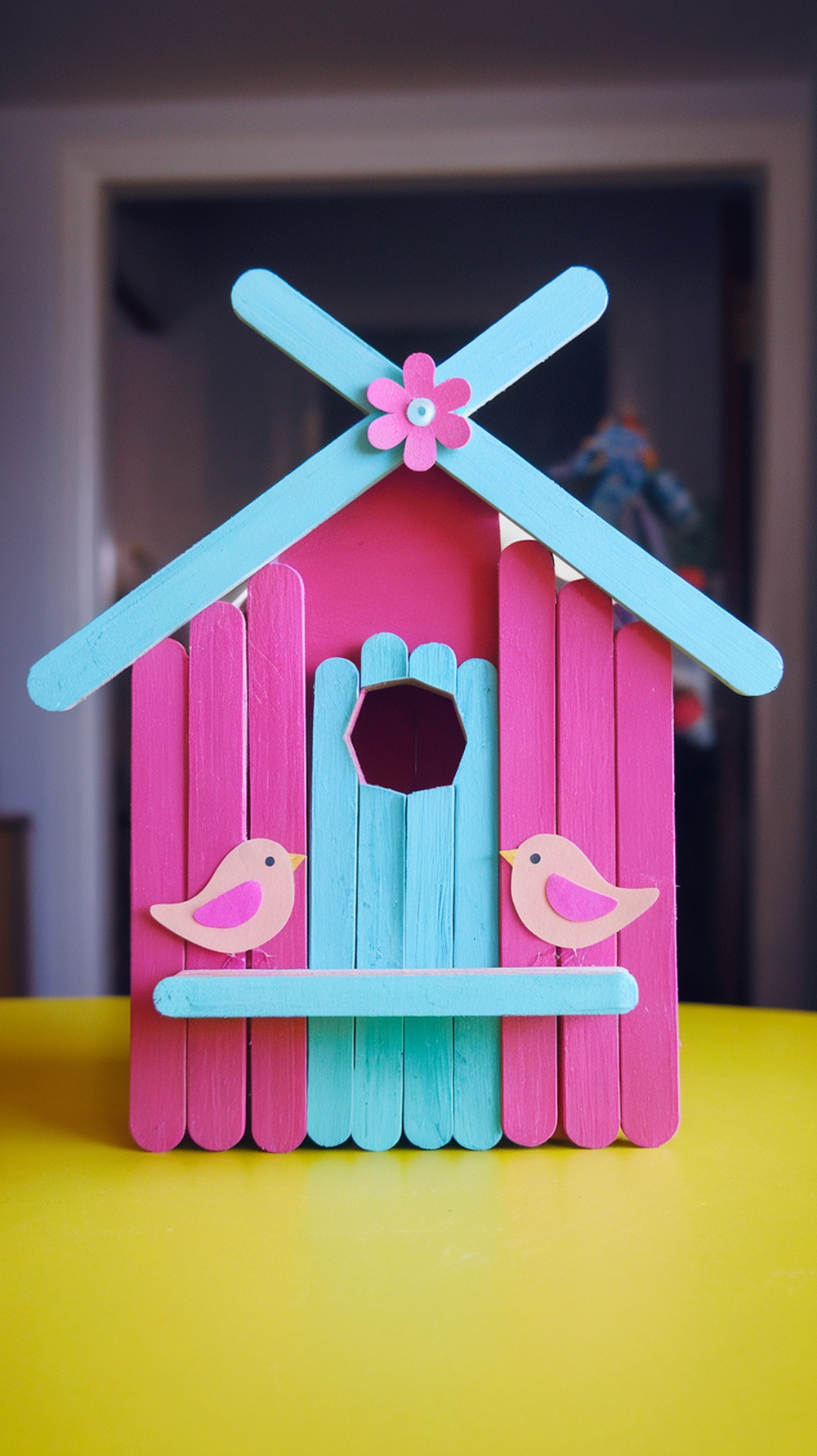 A colorful birdhouse made of popsicle sticks, featuring pink and blue colors, with two little bird decorations.
