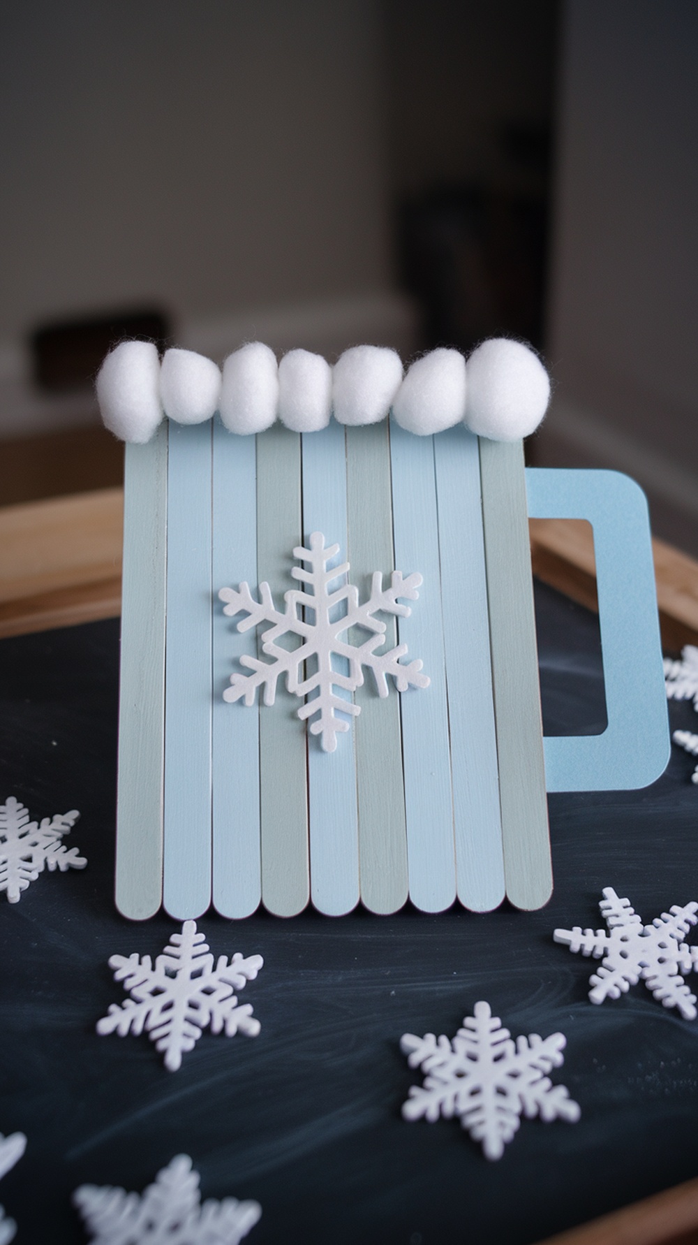 A craft of a winter mug made from popsicle sticks, decorated with cotton balls and a foam snowflake.